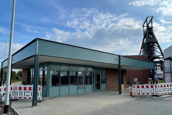 Ein Gebäude in Stahlständerbauweise mit Ziegelstein Mauerwerk. Im Hintergrund ist das Fördergerüst von Schacht Konrad 1 zu sehen.