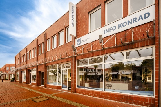 Außendarstellung eines Gebäudes mit roter Backsteinfassade und großen Ladenfenstern in einer Fußgängerzone. Über dem Eingang hängt ein Schild mit der Aufschrift „Info Konrad“.