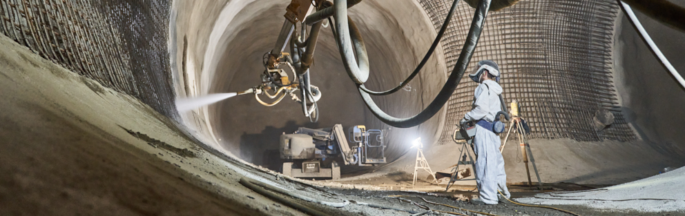 In a brightly lit mine room stands a miner in white work clothes. With a remote control he controls the arm of a large spraying machine