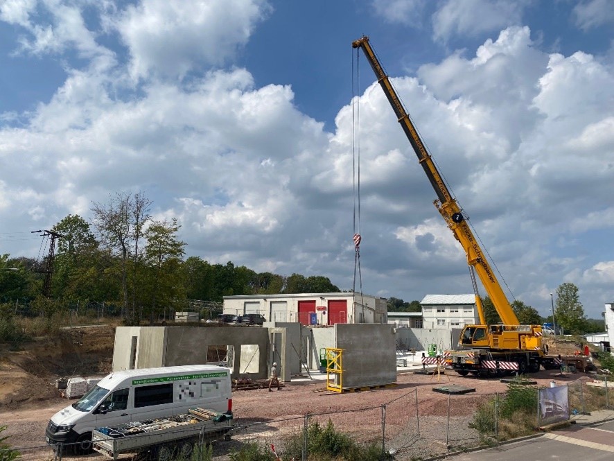 Ein gelber Kran hat eine Betonwand für ein im Bau befindliches Gebäude am Haken. Dahinter ist der Rohbau des Gebäudes zu sehen.