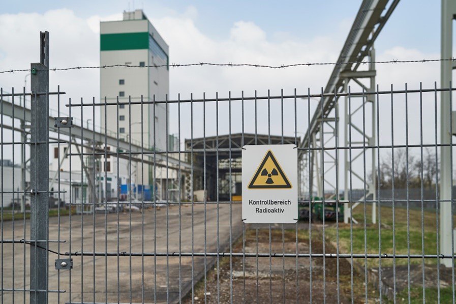 Ein Warnschild zur Markierung eines Kontrollbereichs hängt an einem Drahtzaun. Dahinter sind eine Containerhalle und das Mehrzweckgebäude des Endlagers Morsleben zu sehen. 