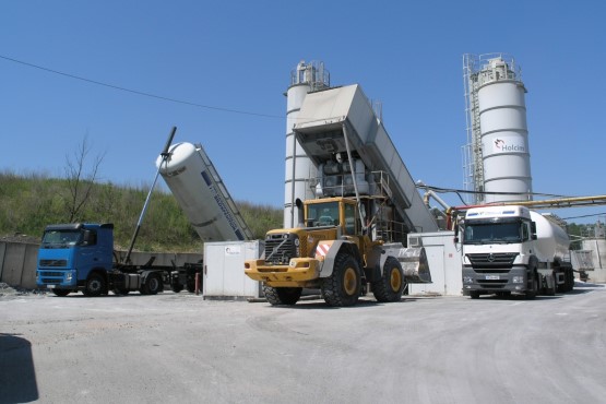 Three vehicles stand in front of a concrete mixing machine
