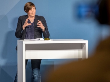 Eine Frau mit dunklen Haaren, einem schwarzen Blazer und Jeans, steht an einem weißen Rednertisch. Ihre Hände formen eine erklärende Geste.